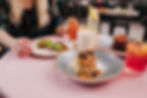 Table setting with pancakes, avocado toast, juice, and a cocktail on a pink tablecloth. A person reaches for food, creating a cozy brunch vibe.
