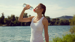 A woman in a white tank top drinks from a water bottle by a lake, with trees and hills in the background, under a clear sky.