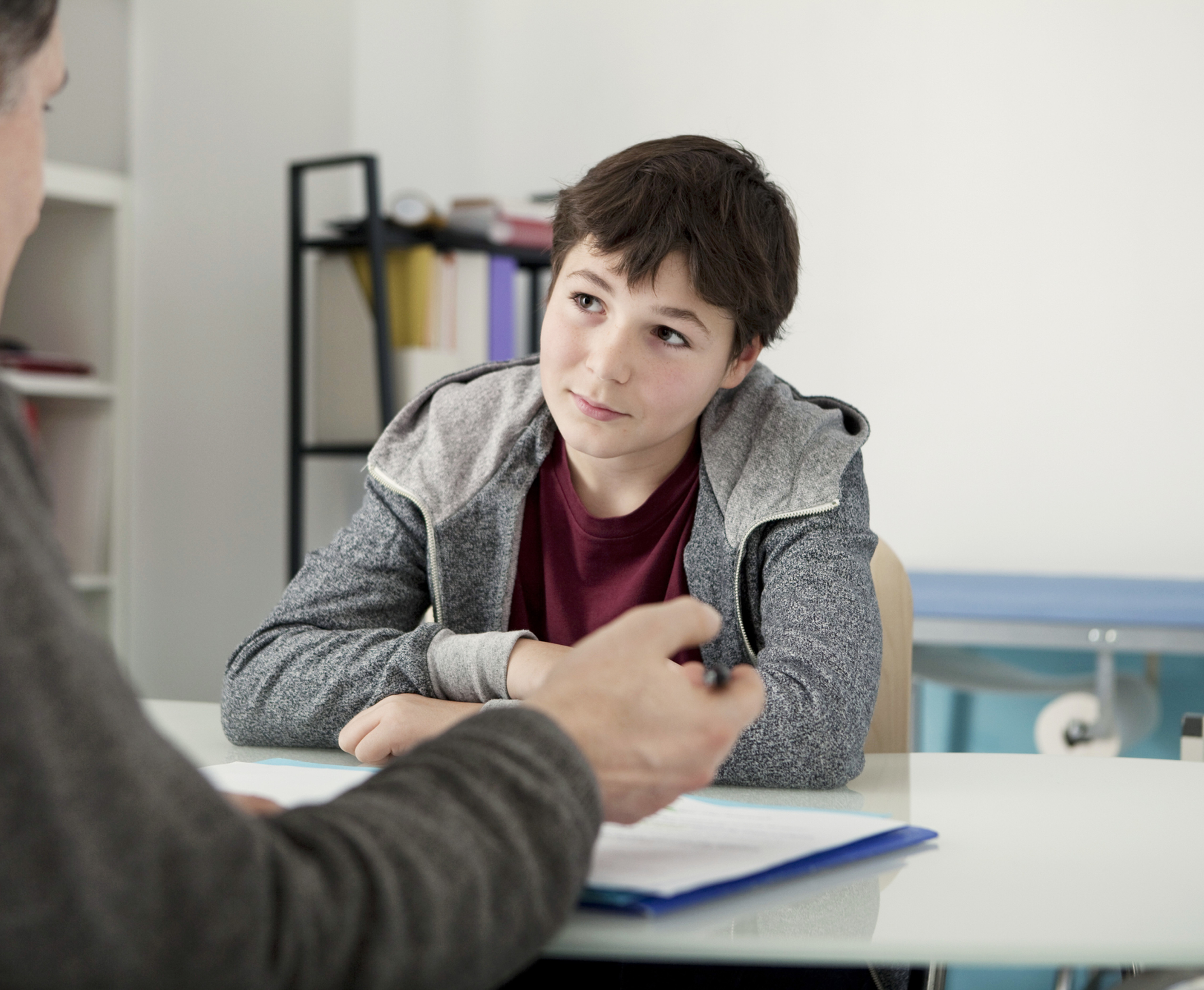 Boy in gray hoodie listens to an adult across a desk in a bright office, appearing attentive during a serious conversation.