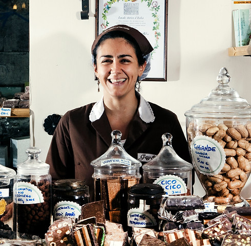 The owner of a dessert shop stands behind her counter of confections smiling.