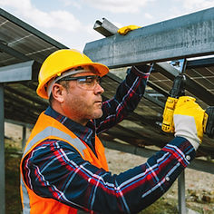 A worker in a hard hat and orange vest uses a drill to install a solar panel