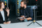 Man with notebook listens to woman gesturing in an office. They're focused, discussing ideas. A smartphone on a tripod records them.