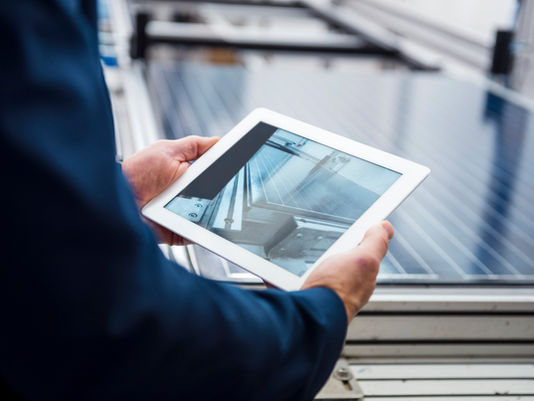 A person in a blue jacket holds a tablet showing solar panels beside a rooftop array.