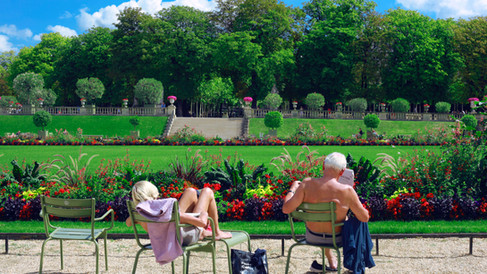 A retired couple relaxing on traditional green chairs in the Jardins de Luxembourg in Paris, on a sunny day with greenery, trees, and flowers all around