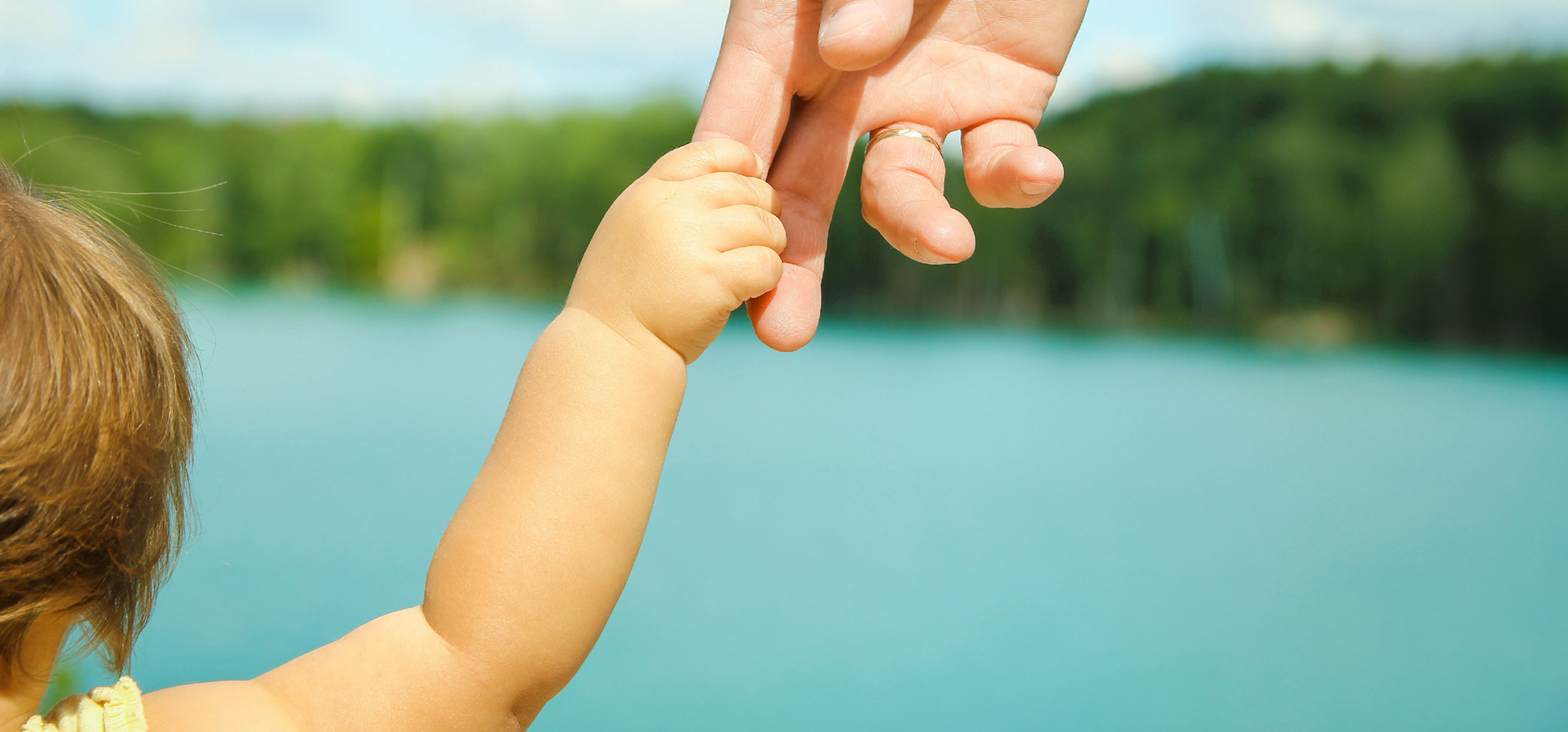 Baby’s hand grips an adult finger against a sunny lake, with blue water, green trees, and sky in the background.