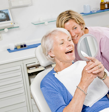 An elderly woman in a dental chair smiles at a handheld mirror, wearing a bib