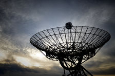 Silhouetted radio telescope dish against a cloudy sunset sky, with the sun glowing behind the metal lattice.