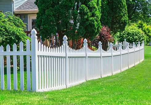 A white vinyl fence running across a yard on a bright summers day with blue sky and trees
