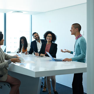 Two adult men and three women at work, in a meeting, in an office with a desk on which is a laptop, several note papers, and pens. They are smiling and look cheerful.