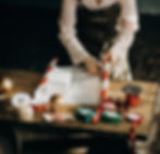 Person wrapping up a gift with red-striped paper on a wooden table, surrounded by ribbons, twine, and gold ornaments.