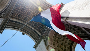 French flag waves under the Arc de Triomphe against a blue sky. Sunlight creates lens flares, highlighting intricate carvings.