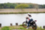 Woman on a motorcycle gazes at a calm lakeside, surrounded by greenery. She's wearing a white shirt and cap, creating a tranquil mood.