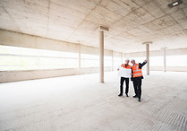 Two men with plan wearing safety vests talking in building under construction