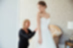 Bride smiles in white gown as woman adjusts her wedding dress.