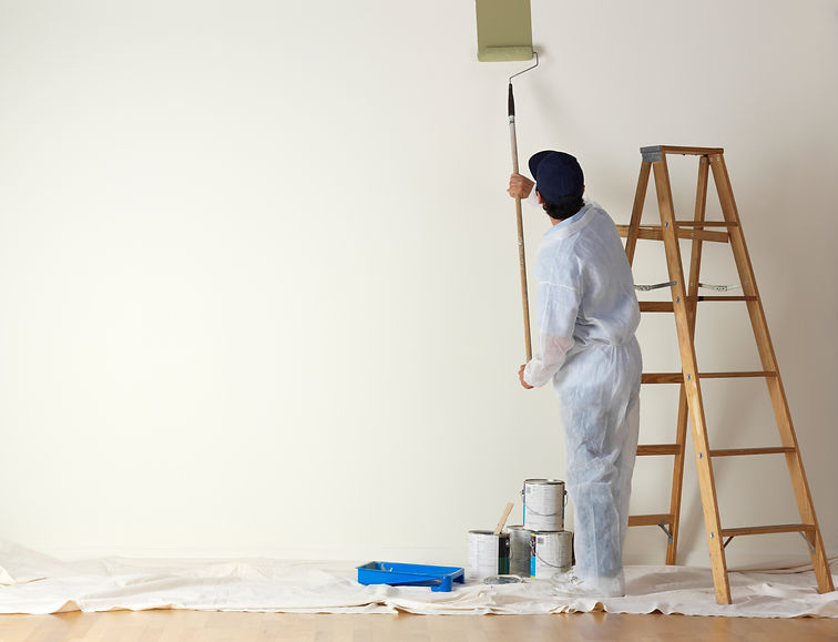 A man dressed in coveralls, uses a paint roller on a long stick to paint a large wall. A