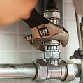 A hand uses a wrench to tighten a metal pipe fitting beneath a tiled surface
