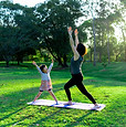 A woman and a child practicing yoga together in a sunny park, surrounded by green grass and trees.
