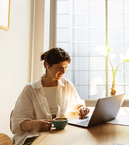 Mulher de cabelo preso de cor castanha usando blusa branca e uma camisa larga por cima com uma xícara de café, mexendo no laptop