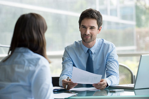 A man in a blue shirt, sitting at a desk, smiles warmly while holding a document