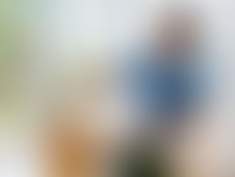 Man painting on canvas indoors, holding a palette, in a bright room with plants. Wears blue shirt, glasses, focused expression.