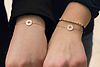 Two wrists wearing delicate rose gold bracelets, each featuring a round compass rose charm with directional N, S, E, W markings