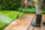 Close up view of a person wearing black rubber boots using a pressure washer lance to spray water onto a stone patio