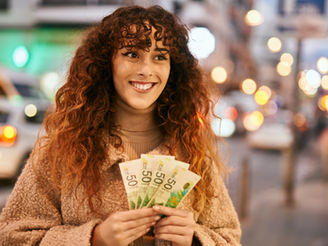 A woman with curly hair smiles happily while holding several 50 currency notes on a bustling city street at night.