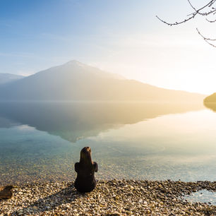 Person sitting by a calm lake, facing a misty mountain under a clear blue sky. Pebble shore and bare branches create a serene mood.