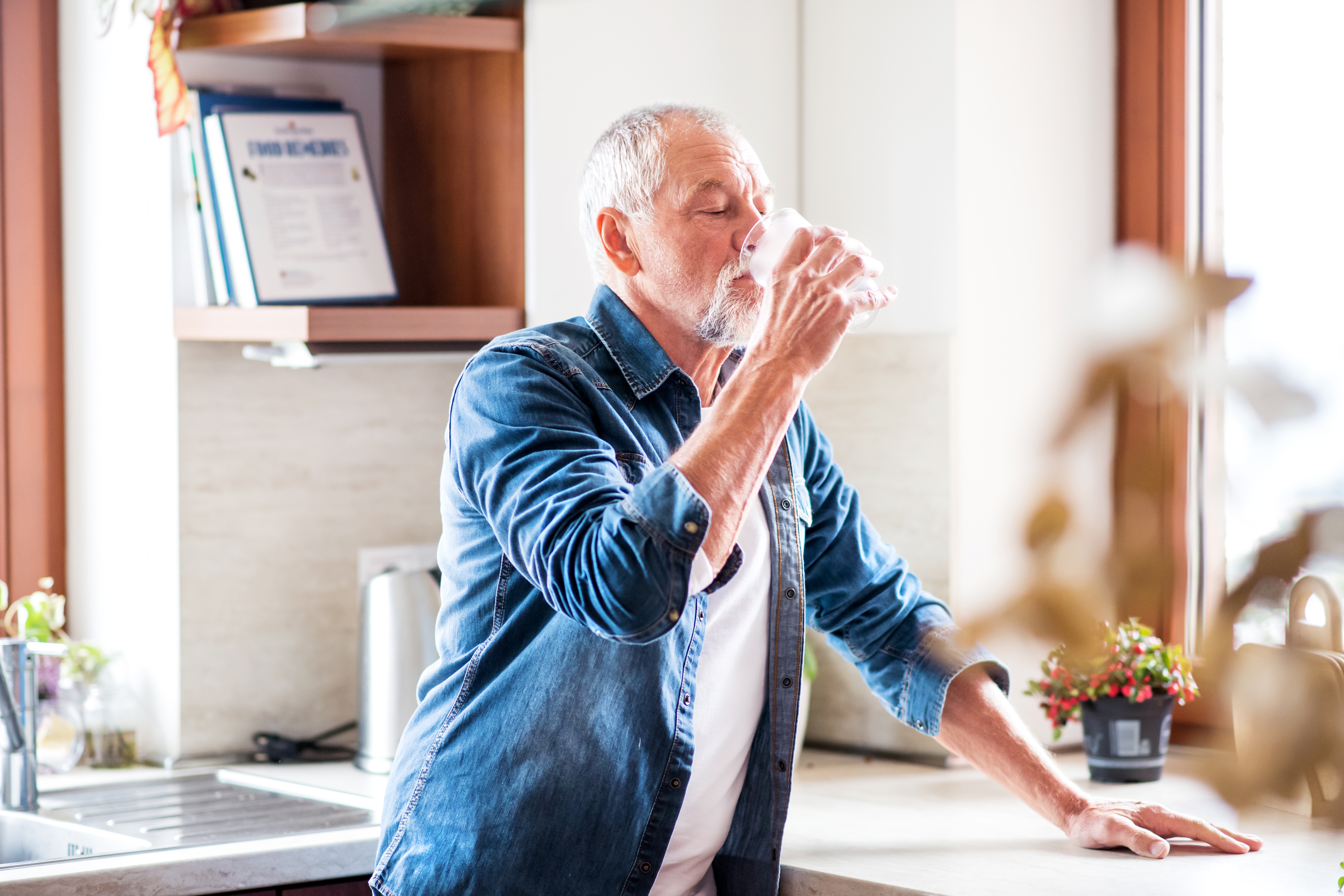 Man with white hair sips water from a glass in a kitchen