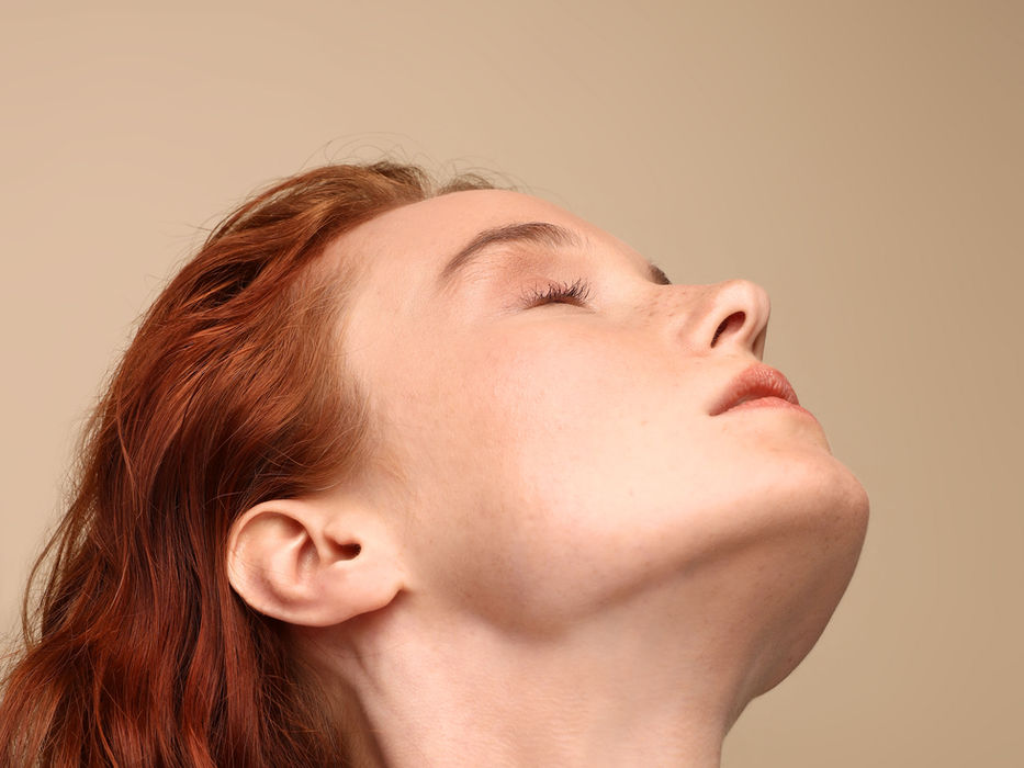 Red-haired woman with eyes closed, tilting head upward, against beige background.