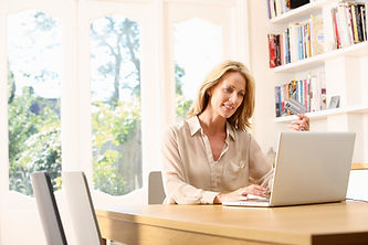 A woman using a laptop while holding a credit card, sitting at a table in a bright, sunny room with bookshelves in the background