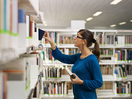 Woman in a library reaches for a book on the shelf, holding another book, with rows of books behind her.