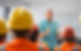 A man in a teal shirt speaks to a group of workers wearing orange safety vests and hard hats