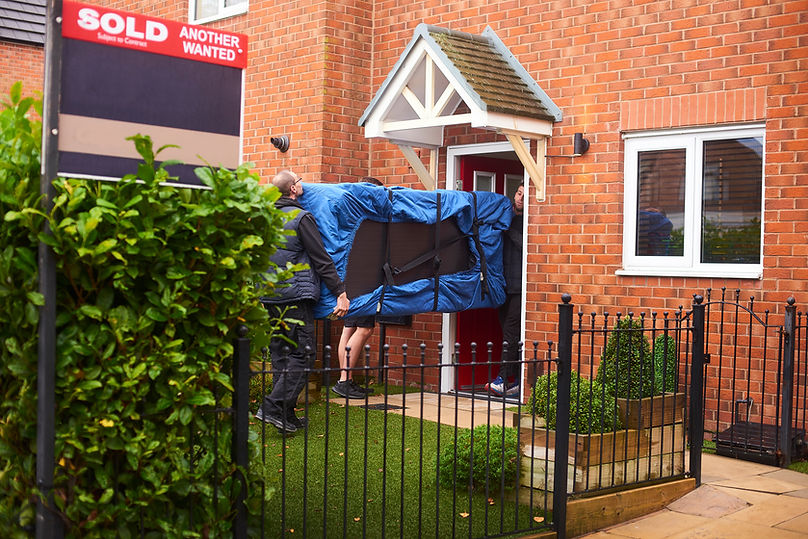Two people carry a blue-covered sofa through a red-brick house