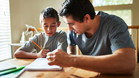 A man and boy sit at a table in a sunlit room, working on homework. The boy writes with a pencil. Both wear gray shirts, focused and calm.