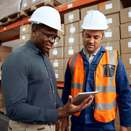 Two warehouse workers in hard hats review a tablet.