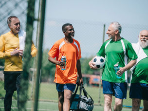 Four men in colorful sports jerseys chat happily on a soccer field. One holds a soccer ball, another a sports bag. Sunny day, fence backdrop.