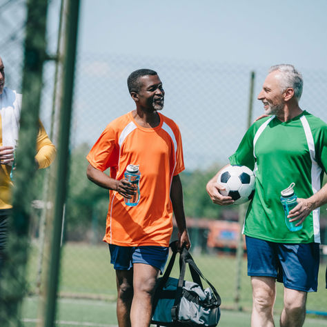 Four men in colorful sports jerseys chat happily on a soccer field. One holds a soccer ball, another a sports bag. Sunny day, fence backdrop.
