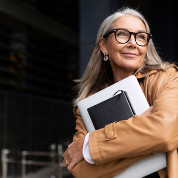Middle-aged woman wearing glasses, smiling while holding a laptop and notebook.