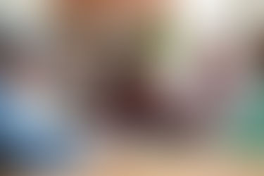 Five seniors smiling while assembling a puzzle at a wooden table in a cozy kitchen with brick walls. Cups are on the table.