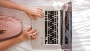 Person in pink pajamas using a laptop on a white bed in soft sunlight, with hands on the keyboard and trackpad.