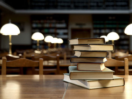 A stack of hardcover books sits on a wooden table in a quiet library reading room, with rows of chairs, tables, and softly glowing desk lamps in the background.