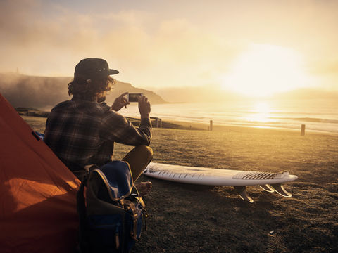 Homme en casquette, assis près d’une tente, photographie un coucher de soleil sur la plage, avec planche de surf.