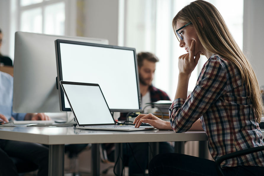 A woman sits at a desk, focused on her laptop, with a notepad and pen beside her