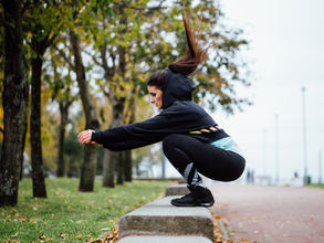 Woman in black hoodie and leggings squats on a concrete ledge in a park. Trees with autumn leaves in the background. Overcast day.