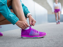 Person in teal jacket ties bright purple running shoe on concrete, another person in pink shorts blurred in the background. Urban setting.