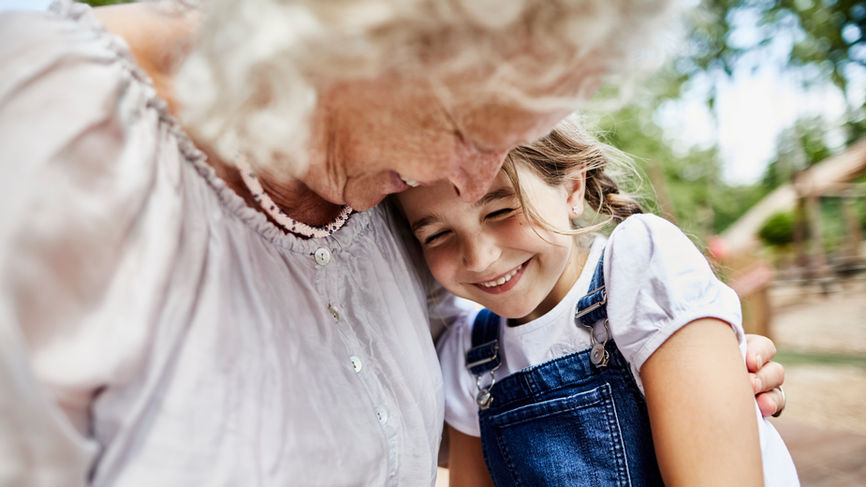 Grandmother with white curly hair and white blouse snuggles her granddaughter outside, with are smiling.