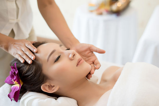 Photograph featuring an Asian woman with closed eyes and a flower in her hair, reclining comfortably. An aesthetic nurse's hands are positioned on her face, indicating the preparation for a chemical peel treatment in an aesthetic salon.