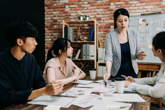Four people in a meeting room discuss charts on a table. A woman in a blazer leads, pointing at papers. Brick wall and shelves in the background.