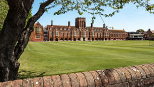 Red brick building with arches and windows stands beyond a lush green lawn. Foreground has a tree and brick wall. Clear blue sky.